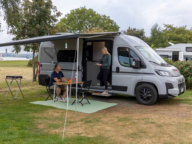 Ricarda steht auf der Stufe das Vans, Lars sitz am Tisch unter der Markise uns schaut in Richtung von Ricarda
