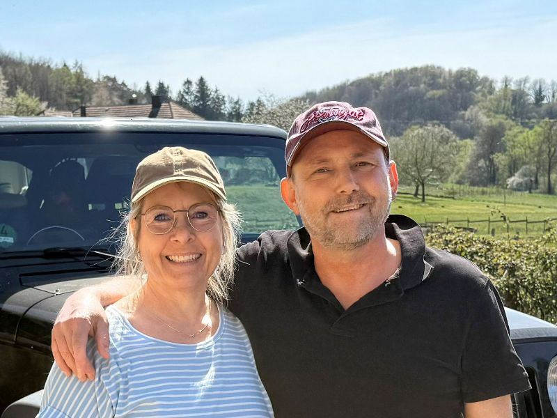 Hertha und Frank stehen vor ihrem schwarzen Jeep, mit grünen Bäumen und Sträuchern im Hintergrund.