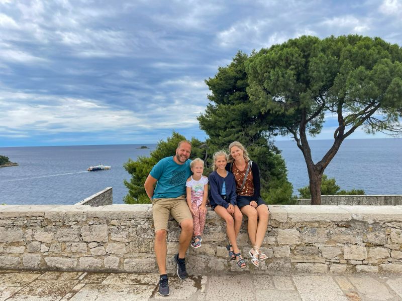 Vater und Mutter mit zwei Töchtern sitzen auf einer Steinmauer mit Pinienbaum und Meer im Hintergrund