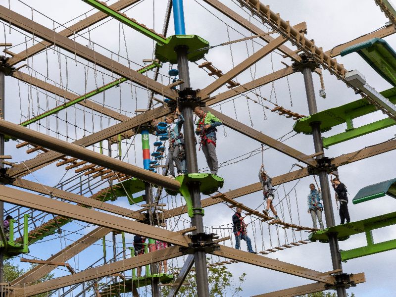 Jens und Leonie stehen weitere vorne im Hinterniss auf dem Klettergarten, während Miriam über ein Seil zu ihnen läuft. Petra wartet.