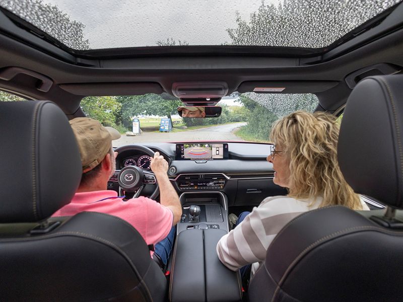 Frank und Herta sitzen im Mazda CX-60 mit Panoramadach und fahren auf den Campingplatz auf. 