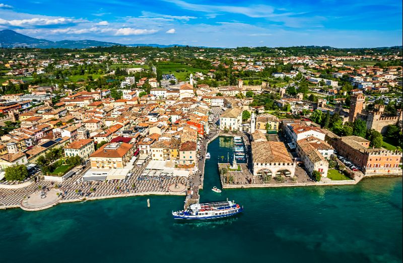 Luftaufnahme der Altstadt von Lazise mit Hafen, Uferpromenade und Blick über die umliegende Landschaft am Gardasee.