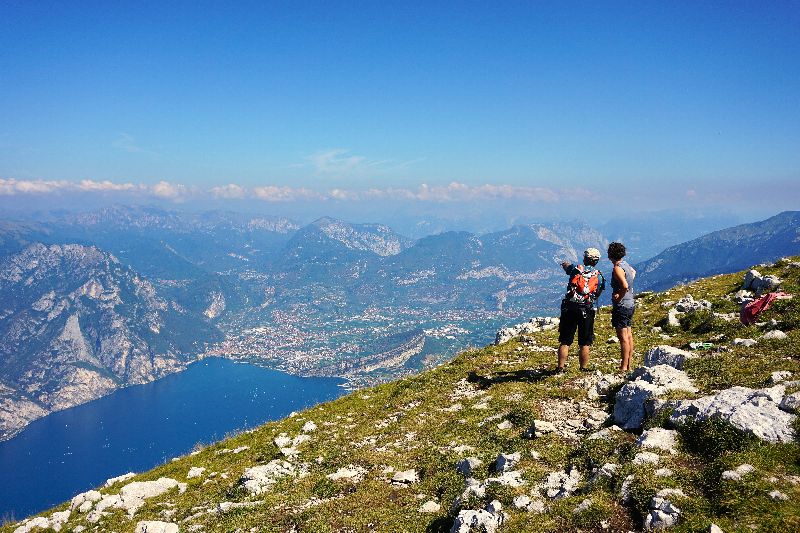 Zwei Wanderer stehen auf dem Monte Baldo und blicken über den Gardasee und die umliegende Berglandschaft.