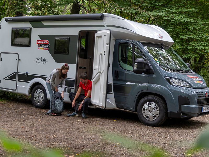 Maja und Felix im Wald vor ihrem Camper mit Wanderschuhen und Rucksack