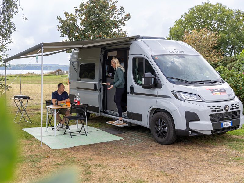 Lars sitzt unter dem Vorzelt des Vans und Ricarda steht auf der Stufe vom Etrusco CV 600 SB