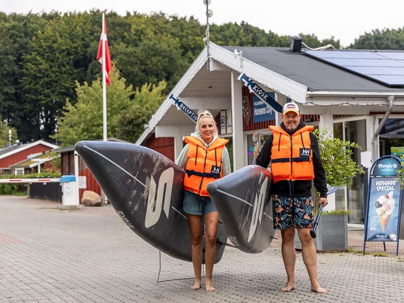 Ricarda und Lars sind mit SUP-Brettern und Schwimmwesten ausgestettet und stehen vor dem Hauptgebäude des Campingplatzes