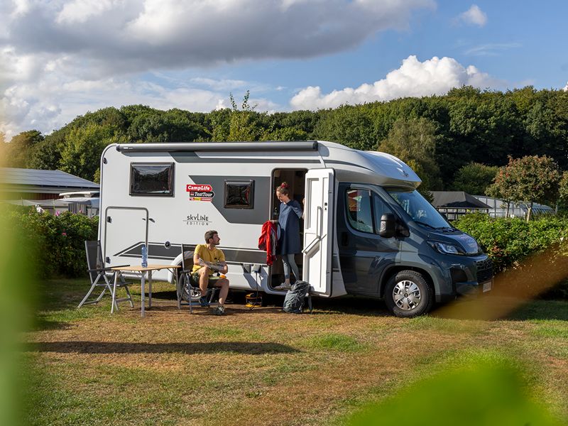 Felix sitzt auf einem Campingstuhl vor dem Camper, Maja steht an der Tür um aus dem Bürstner zu steigen