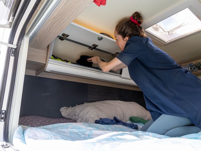 Maja holt ein Kleidungsstück aus dem Fach oberhalb des Bettes.