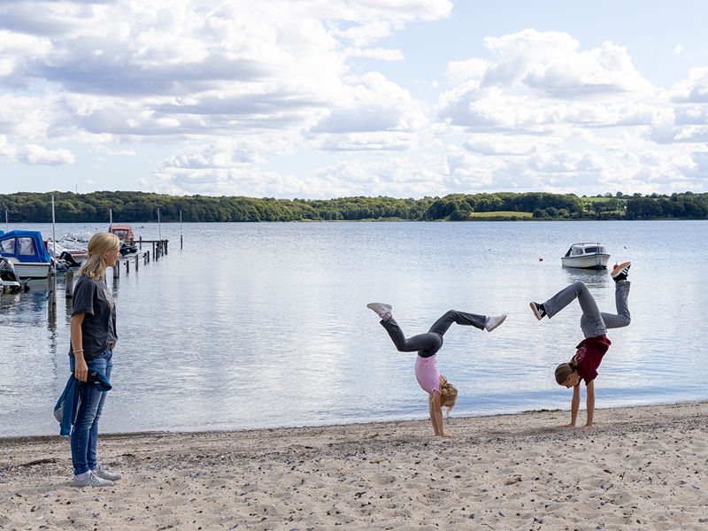Leonie und Miriam machen einen Handstand am Strand und Petra schaut lachend zu. Ein kleines Boot treibt im Hintergrund
