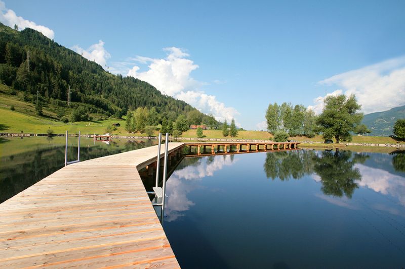 Ein Holzsteg an einem ruhigen Bergsee beim Sportcamp Woferlgut, mit grünen Hügeln und blauem Himmel im Hintergrund. 