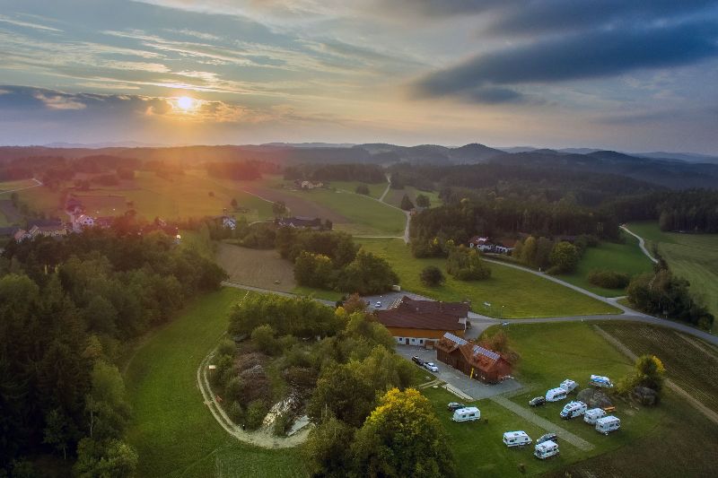 Luftaufnahme des Campingplatzes Waldviertel bei Sonnenuntergang, mit Stellplätzen, hügeliger Landschaft und Wäldern in Österreich